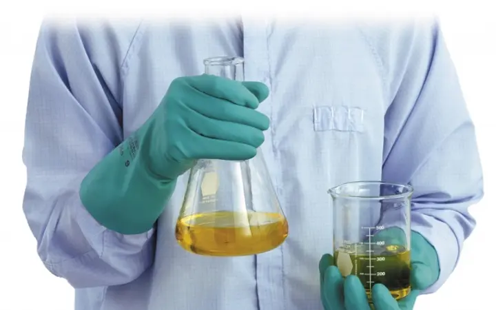 Kỹ thuật viên đeo găng tay nitrile chống hóa chất màu xanh lá đang cầm bình tam giác và cốc đong chứa dung dịch trong phòng thí nghiệm