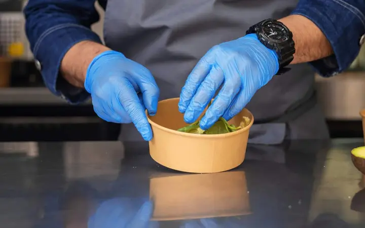 Nhân viên bếp đeo găng tay nitrile xanh trộn salad trong hộp giấy